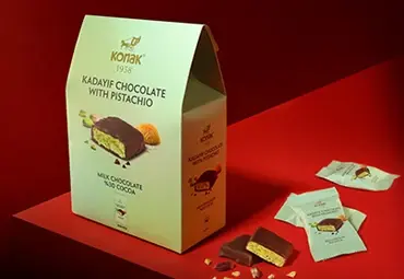 Konak Kadayif Chocolate with Pistachio gift box displayed with unwrapped bars and crumbs on a red surface.